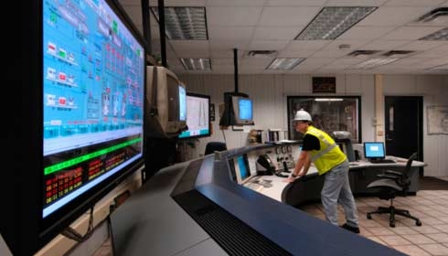 Service-Techniker mit Helm betrachtet Computerbildschirme in Bedienraum, Gebäudemanagementsoftware.