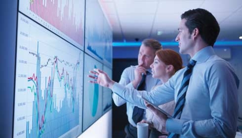 Three business people discuss graphs on screen in meeting room