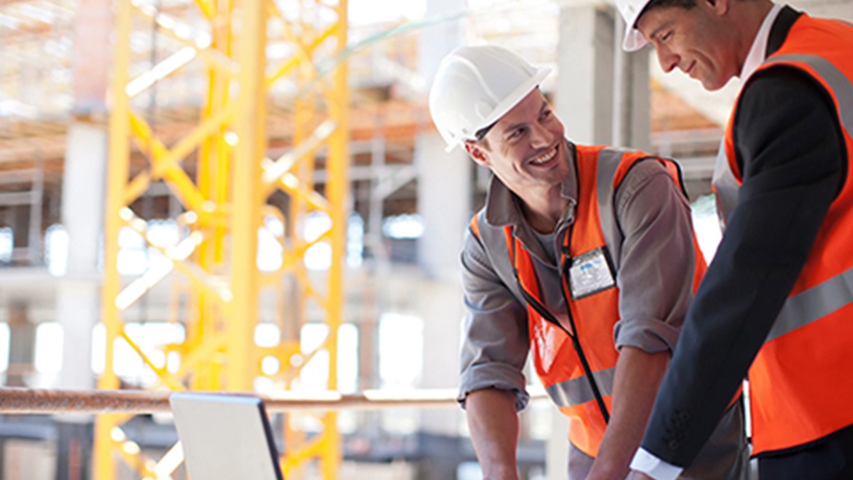 two men in hardhats looking at laptop and smiling