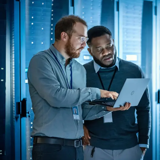 A person holding a computer and another person standing in a server room