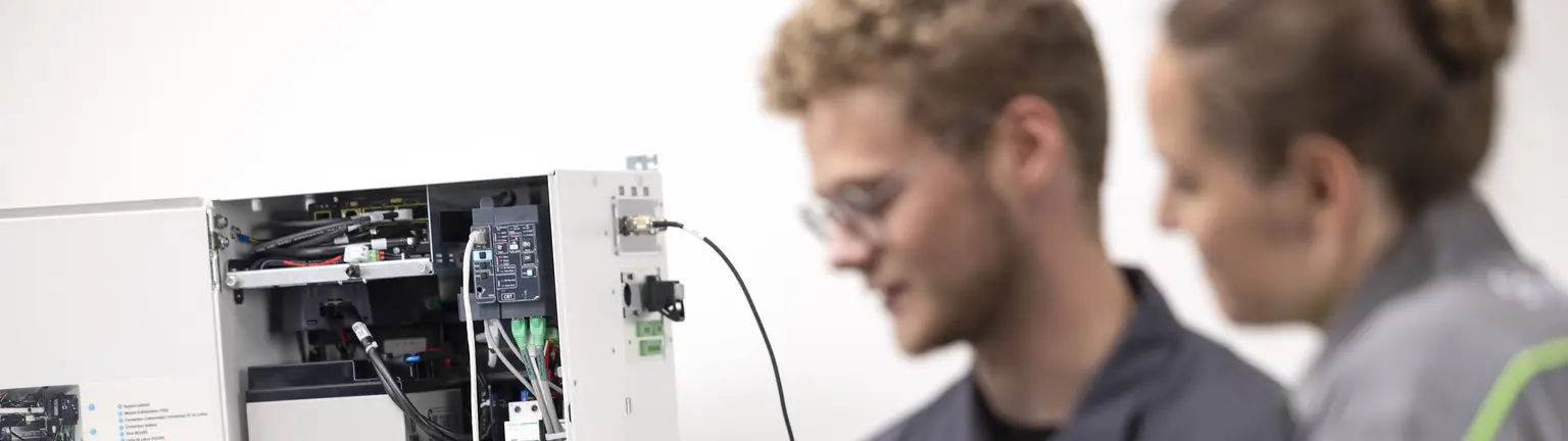 Two technicians inspect an open electrical panel; one holds a laptop for diagnostics