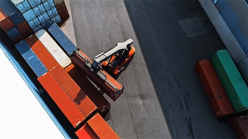 container handler carrying a large red cargo container in a shipyard terminal, powered by an optimized electrical distribution system for sustainable port infrastructure