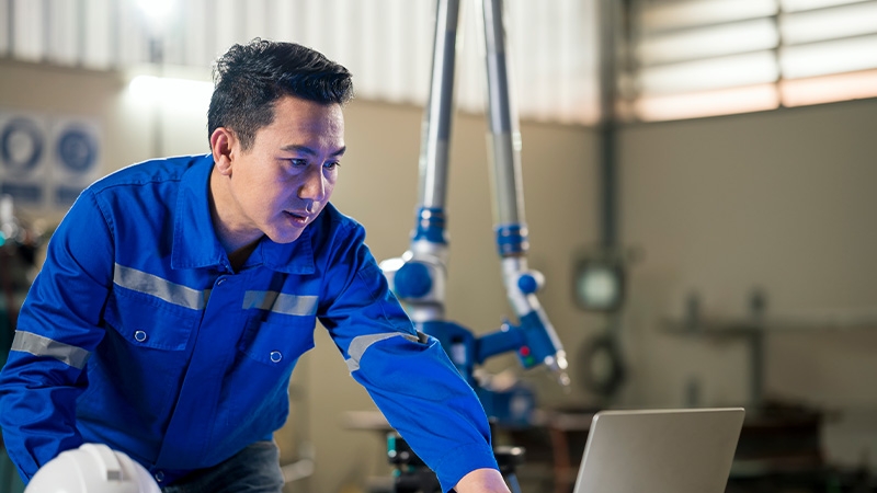 Asian Male Engineer using laptop in industrial robotic arm. Manufacturing process improvement ideas.