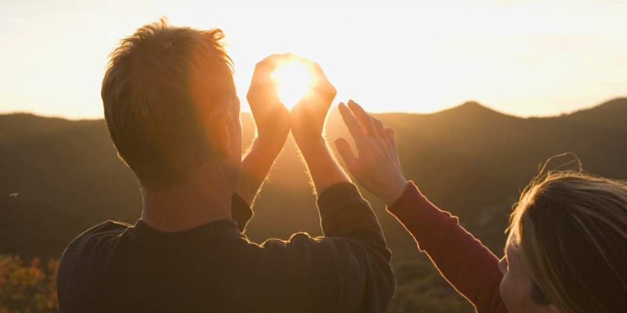 Un homme et une femme regardent le coucher de soleil. L'homme fait comme s'il tenait le soleil dans ses mains.