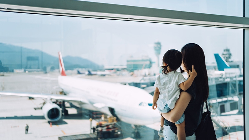 Joyful young Asian mother embracing cute little daughter looking at airplane through window at the airport while waiting for departure
