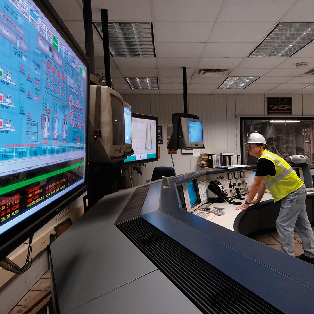 Technician in hard hat monitoring computer screens in control room, facility management software.
