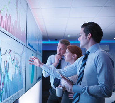 Three business people discuss graphs on screen in meeting room