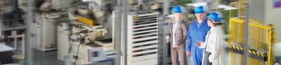 Business people in hard-hats walking on factory floor with lots of machines, power management, plant solutions.