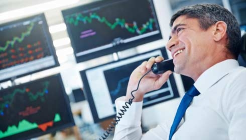 Profile shot of a laughing businessman talking on the phone while sitting in front of monitors displaying financial information