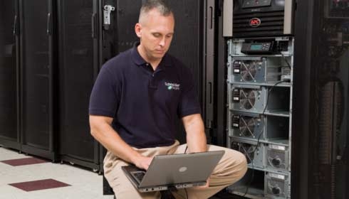 A man squatting in front of a server console with his laptop.