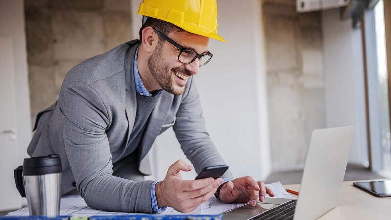 Young smiling architect in suit with helmet on head holding smart phone and using laptop while leaning on table in building in construction process.