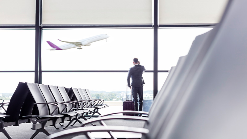 Businessman with suitcase talking on cell phone at smart airport departure area window  