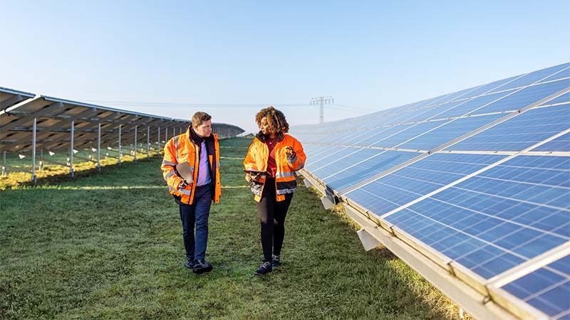 A man and woman in orange jackets walk in front of solar panels, promoting renewable energy for electric utilities.