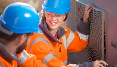 A female air conditioning technician with her supervisor on a roof top of an office block . They are performing routine servicing of the air conditioning units , and are wearing safety equipment .