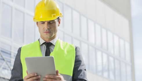 Young male engineer in reflector-vest and hardhat using digital tablet outside industry