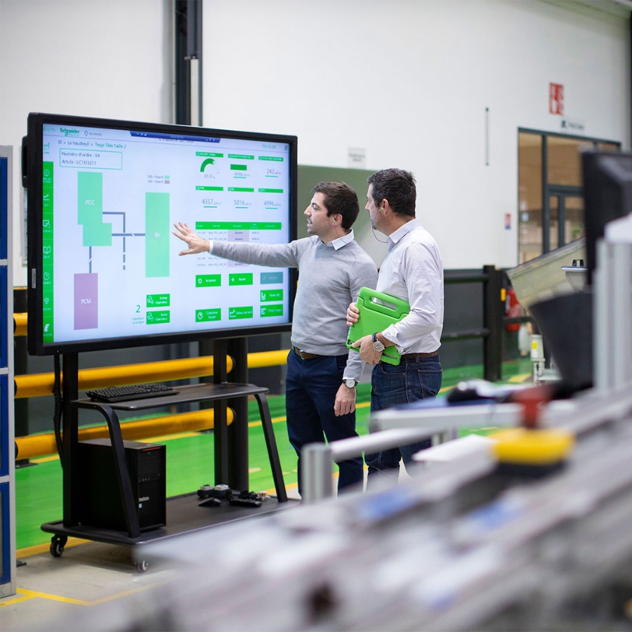 Two male employees discussing a presentation slide which is on a big screen inside a factory